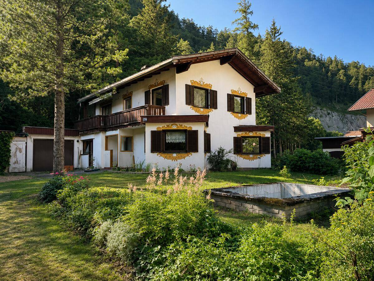 Traditionelles Haus mit weißer Fassade, braunen Fensterläden und einem grünen Garten vor bewaldeten Bergen.