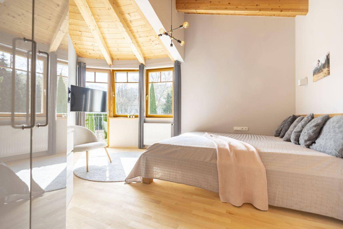 Helles Schlafzimmer mit Holzbalkendecke, großen Fenstern und Blick ins Grüne.