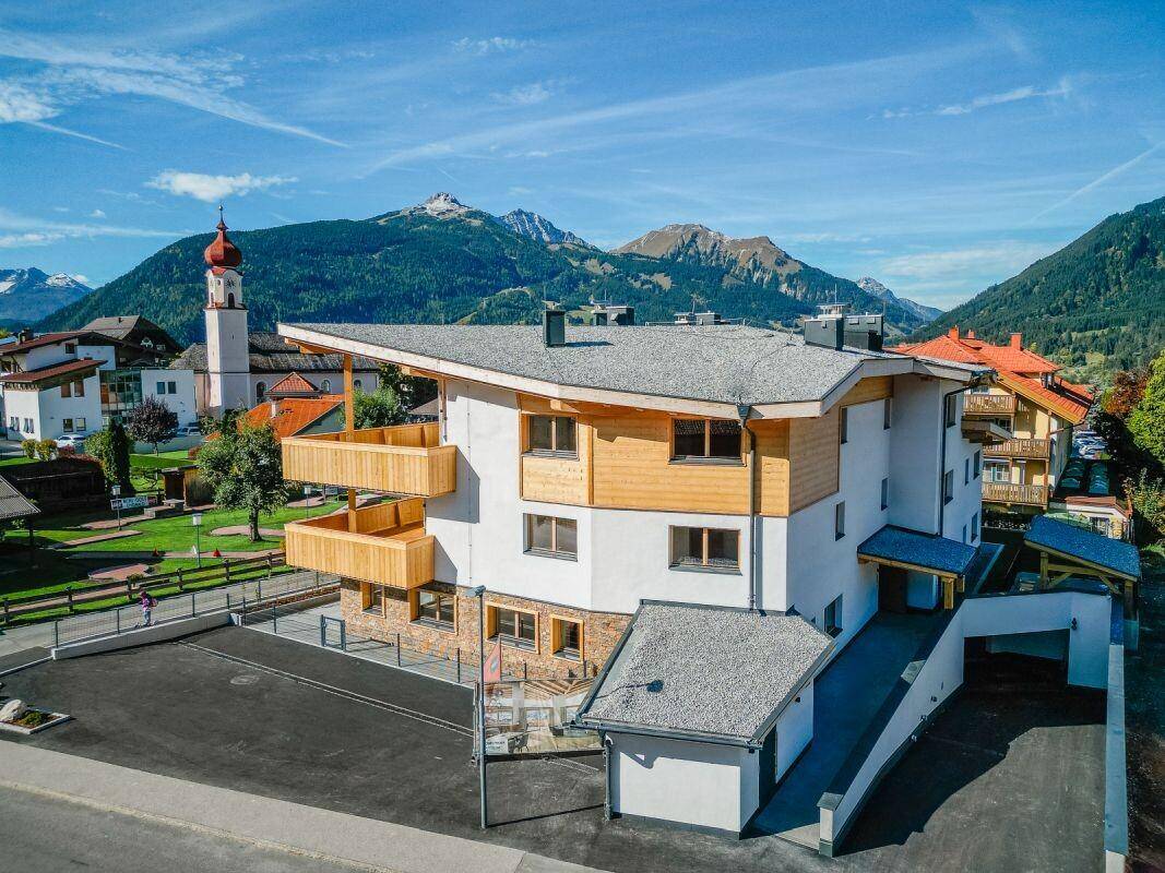 Luftaufnahme des modernen Gebäudes in einer malerischen Berglandschaft mit Kirchturm im Hintergrund.