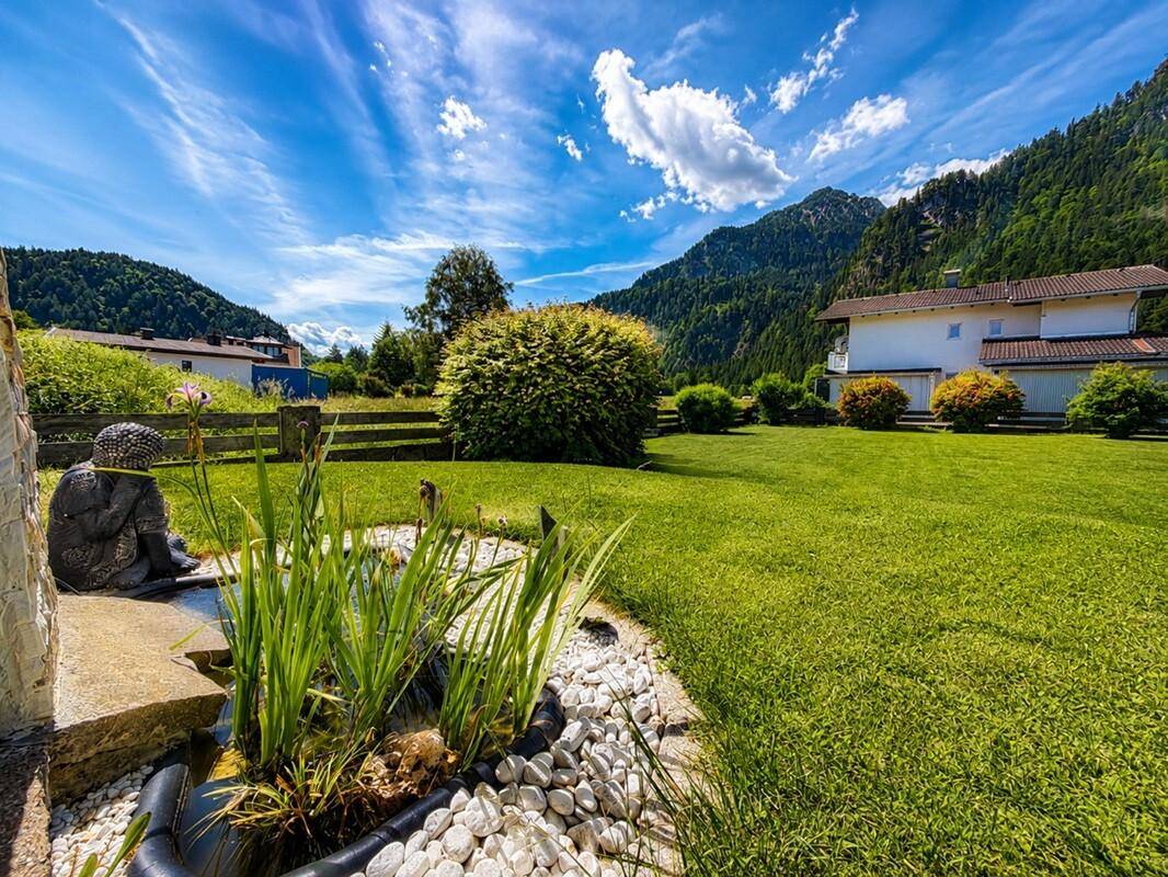 Gepflegter Garten mit Teich, Rasenfläche und Bergblick unter blauem Himmel.