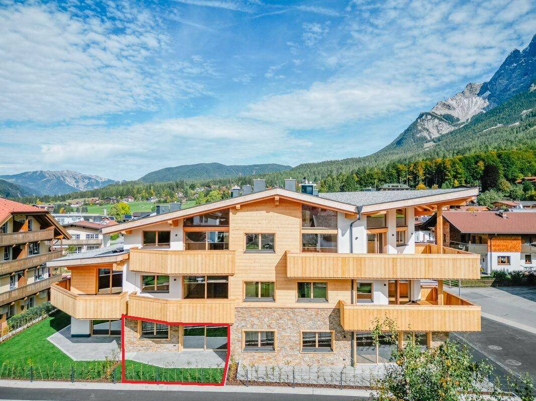 Luftaufnahme des Neubaus mit Balkonen und einer hervorgehobenen Erdgeschossterrasse vor Bergkulisse.