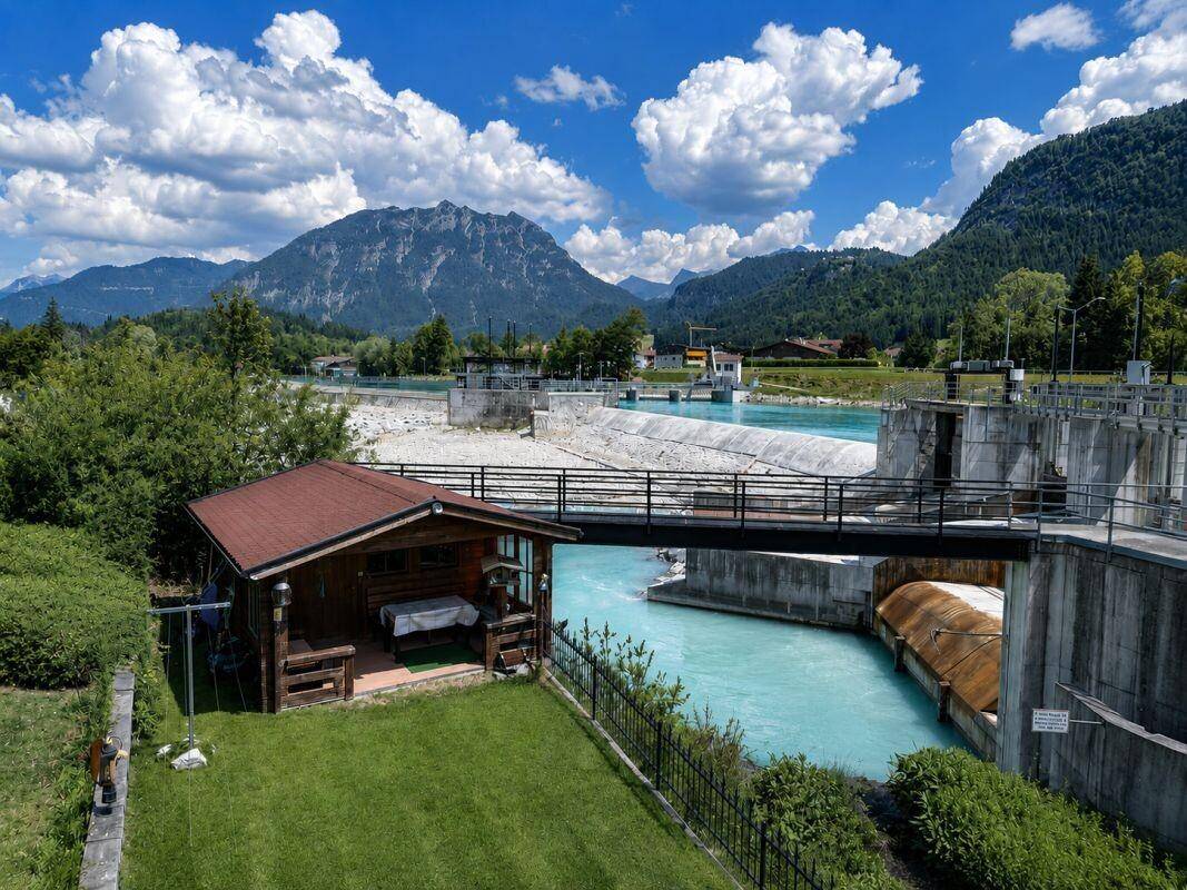 Gartenhütte am Flussufer mit Brücke und Bergkulisse unter blauem Himmel.