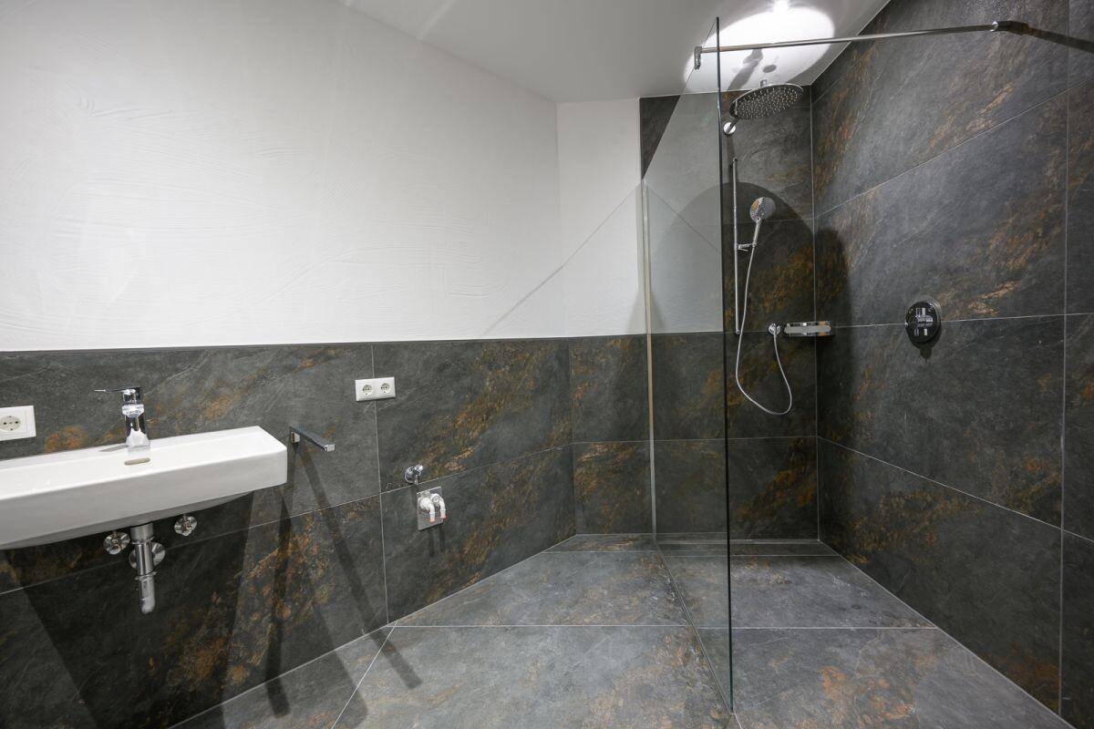 Modernes Badezimmer mit dunklen Fliesen, weißem Waschbecken und großer begehbarer Dusche.