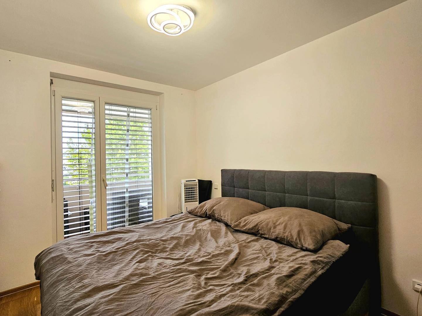 Schlafzimmer mit großem Bett, Fenster mit Jalousien und Zugang zu einem Balkon.