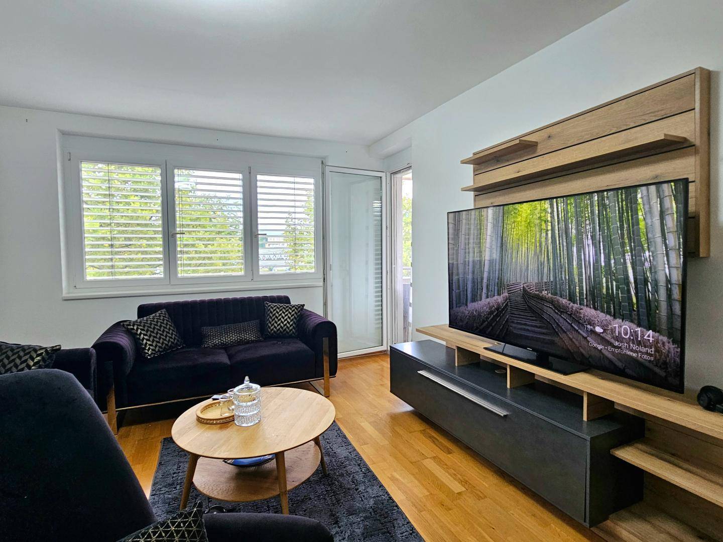 Gemütliches Wohnzimmer mit dunklem Sofa, großem Fernseher und Zugang zu einem Balkon.