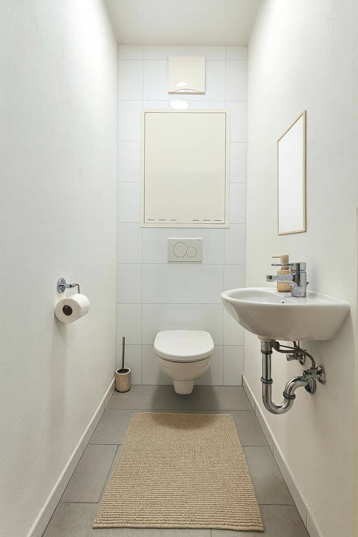 Schlichtes Gäste-WC mit wandhängender Toilette, kleinem Waschbecken und hellen Fliesen.