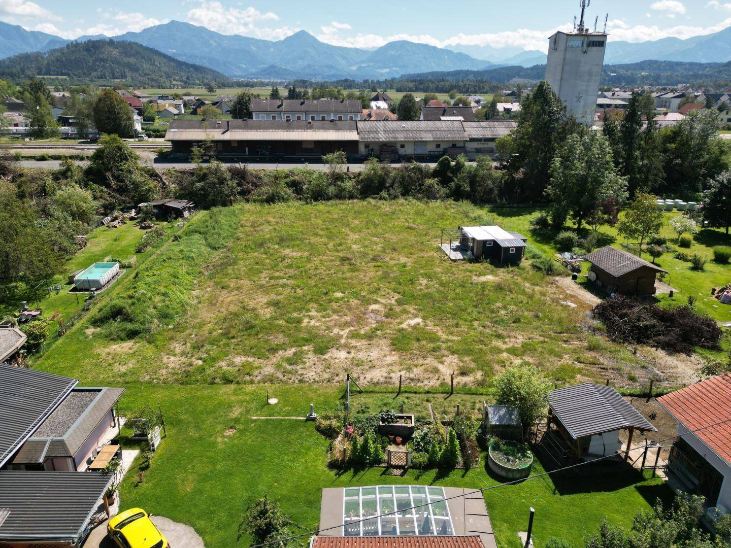 Luftaufnahme des Grundstücks mit kleiner Hütte, angrenzendem Garten mit Gewächshaus und Bergpanorama.