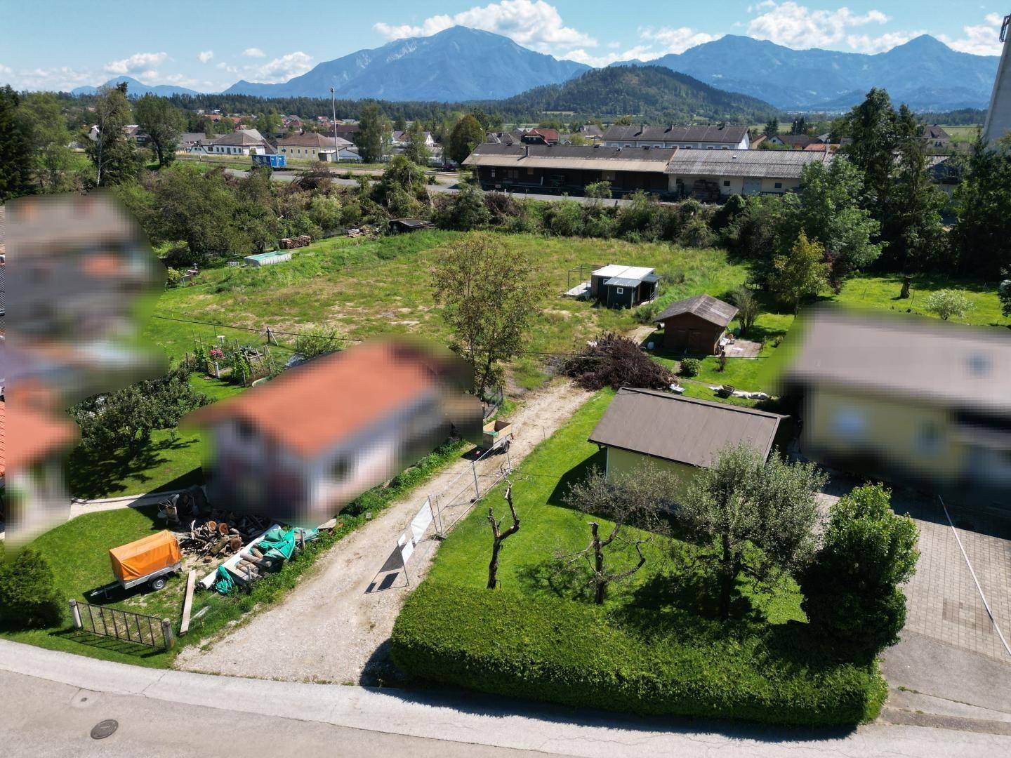 Luftaufnahme eines Grundstücks mit unscharfen Nachbargebäuden, Hecken und Bergkulisse im Hintergrund.
