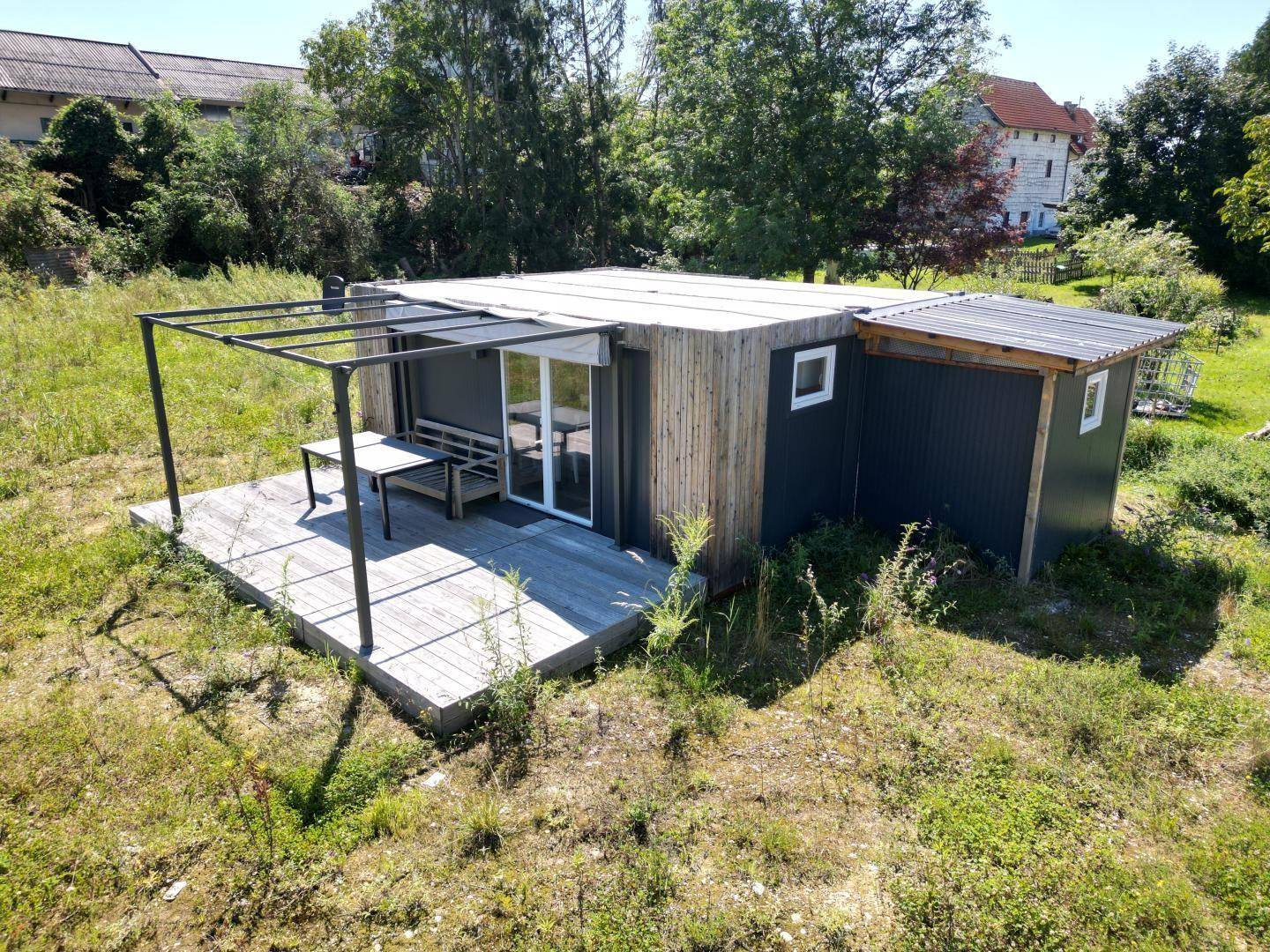 Kleine Hütte mit Holzterrasse und Pergola-Rahmen, umgeben von grüner Wiese.