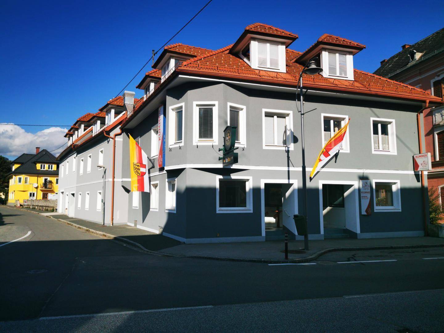 Ansicht des grauen Gebäudes mit rotem Dach und Dachgauben an einer sonnigen Straßenecke mit blauem Himmel.