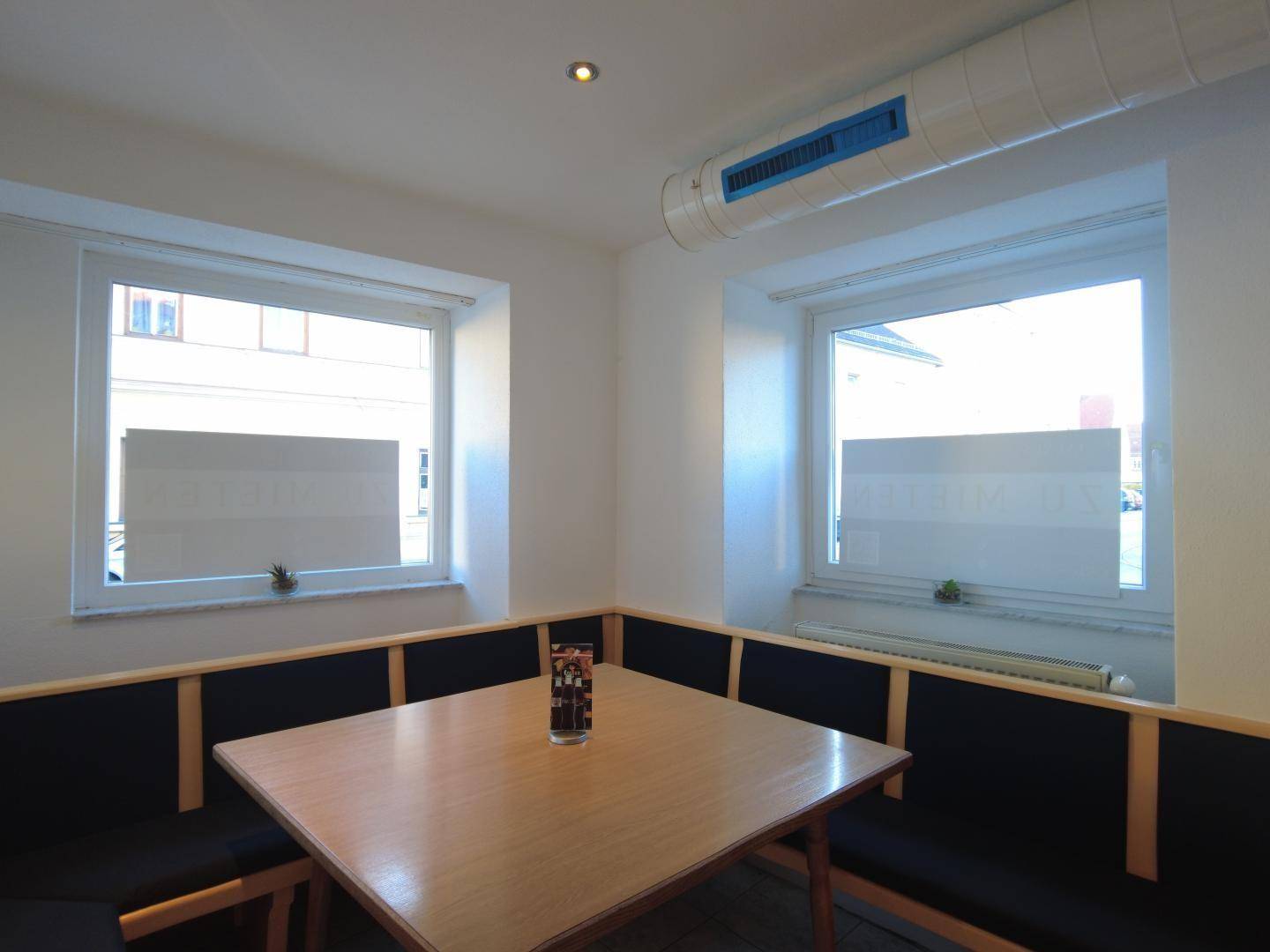 Gemütliche Sitzecke mit gepolsterten Bänken, Holztisch und zwei Fenstern, die Tageslicht hereinlassen.