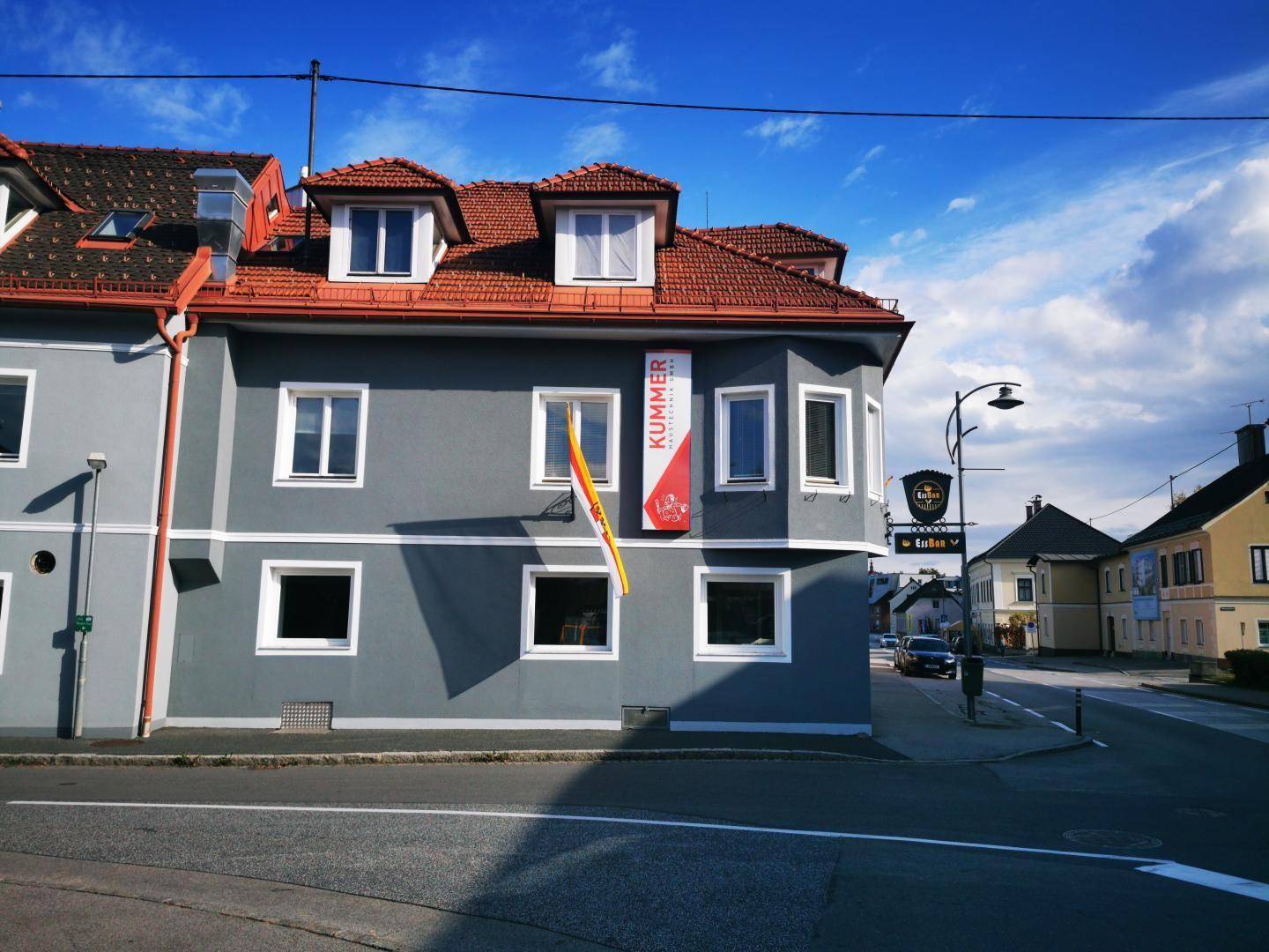 Seitenansicht des grauen Gebäudes mit rotem Dach, Dachgauben und einem Werbebanner an der Fassade.