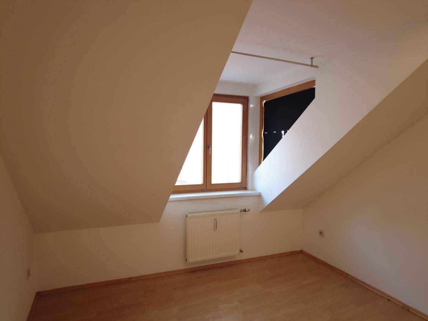 Helles Zimmer im Dachgeschoss mit Holzboden und Fenster, das viel Tageslicht hereinlässt.