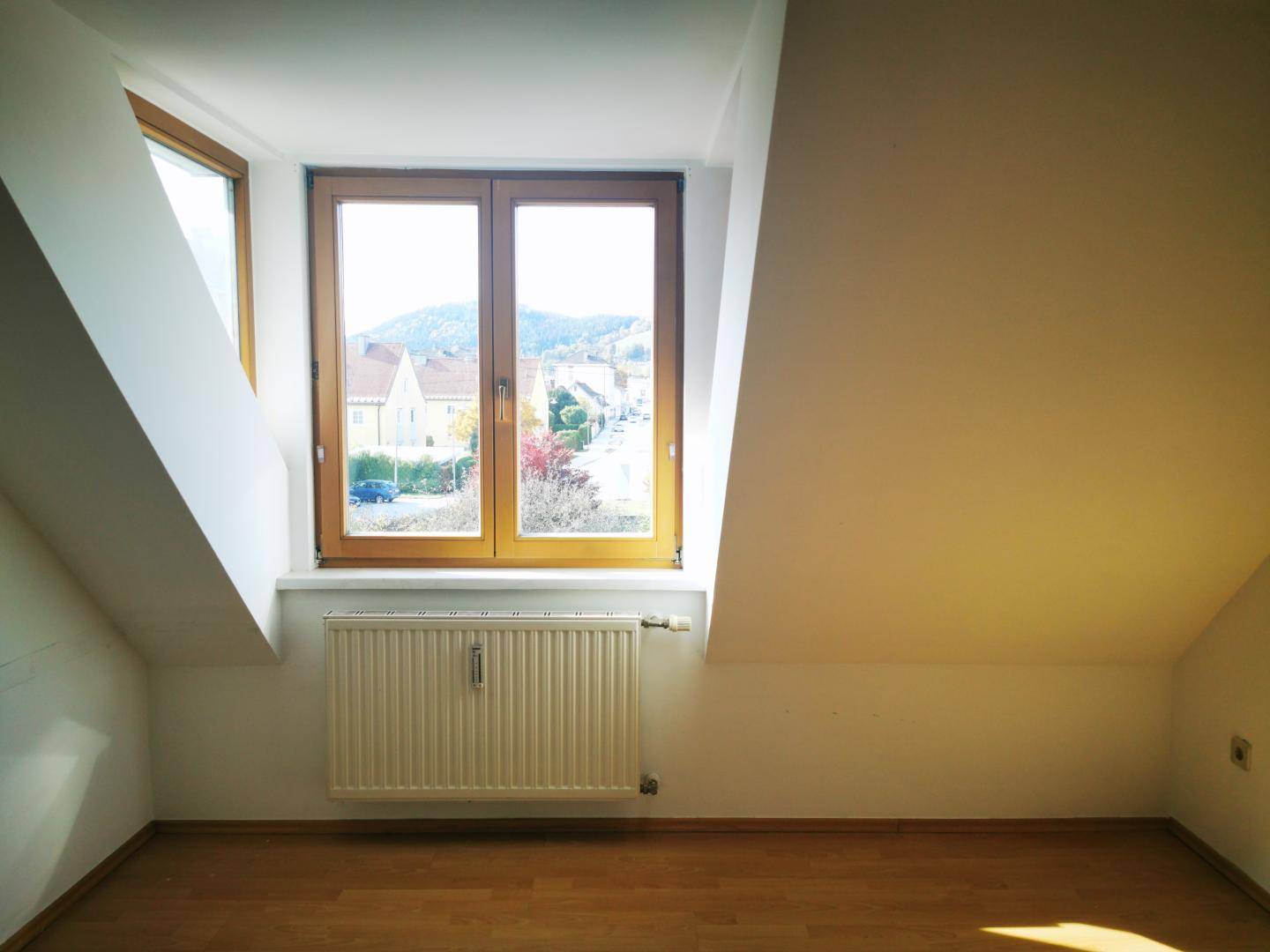 Helles Zimmer im Dachgeschoss mit Holzboden und großem Fenster mit Blick auf die Umgebung.