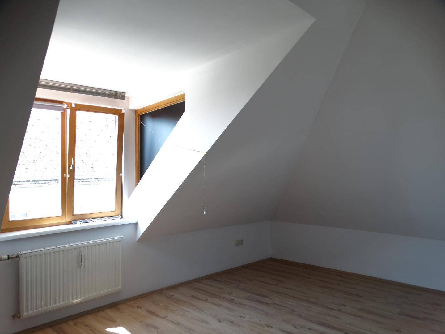 Helles Zimmer im Dachgeschoss mit Holzboden und Fenster, das viel Tageslicht hereinlässt.