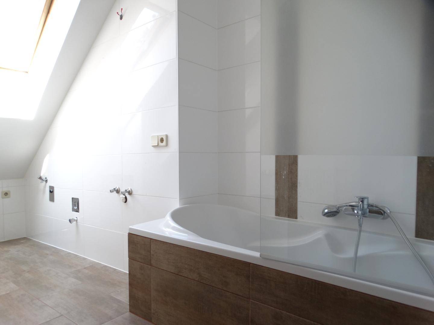 Modern gefliestes Badezimmer im Dachgeschoss mit Badewannenbereich und vorbereiteten Anschlüssen.