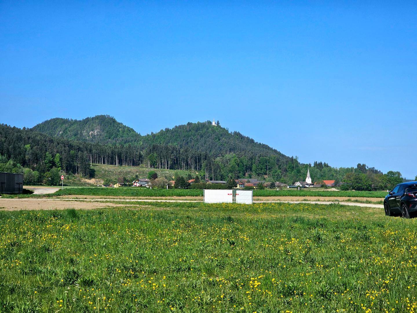 Offenes Feld mit Blick auf ein malerisches Dorf und bewaldete Hügel unter klarem blauem Himmel.