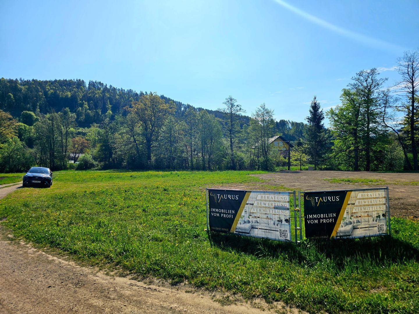 Grüne Wiese mit Immobilienwerbetafeln und bewaldeten Hügeln unter einem klaren blauen Himmel.