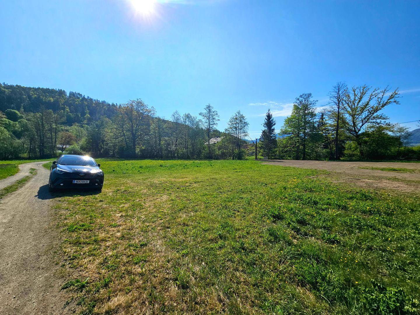 Unbefestigter Weg neben einer grünen Wiese mit einem Auto und bewaldeten Hügeln im Hintergrund.