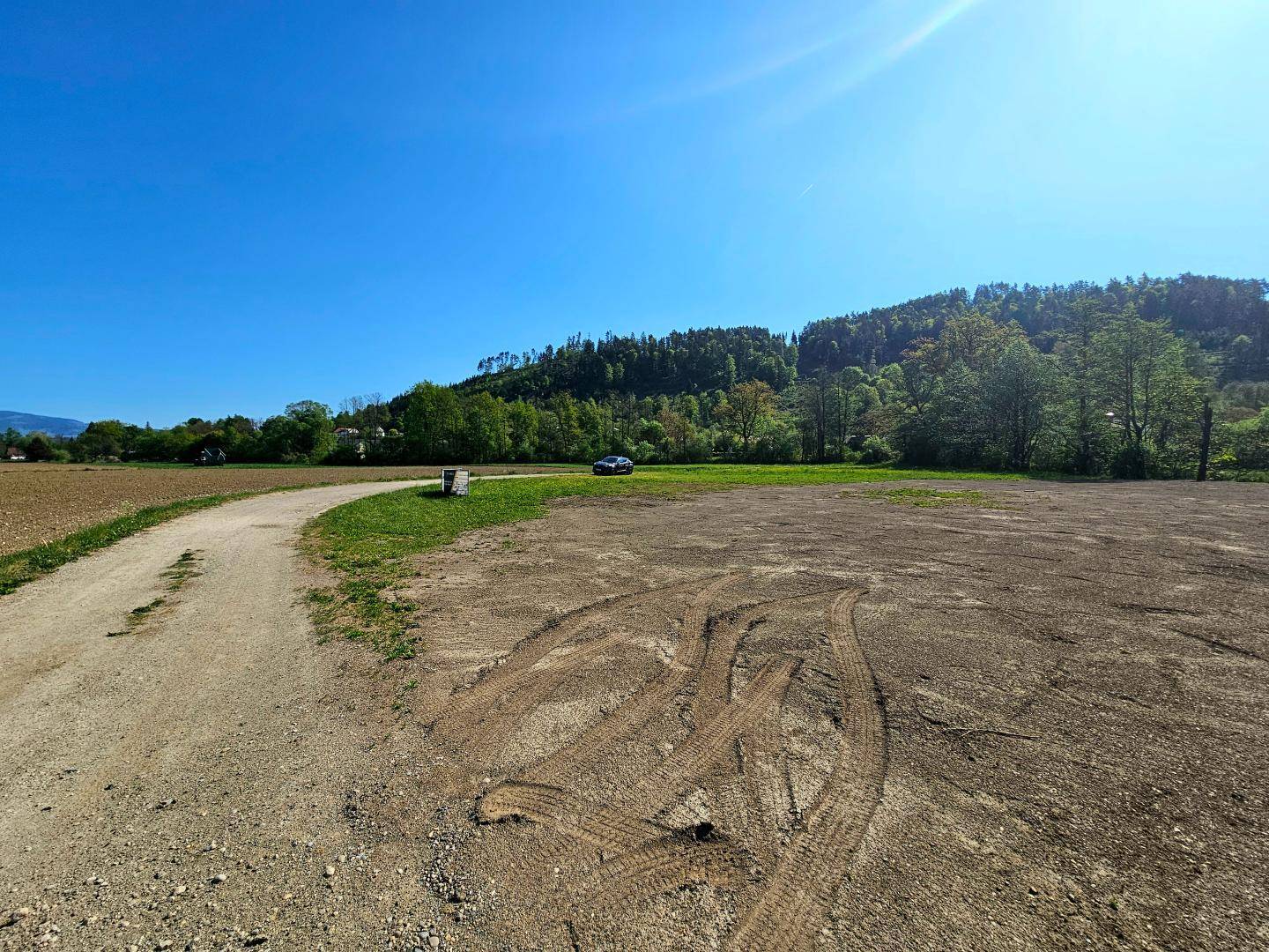 Unbefestigter Weg führt durch ein freigelegtes Grundstück mit bewaldeten Hügeln im Hintergrund.