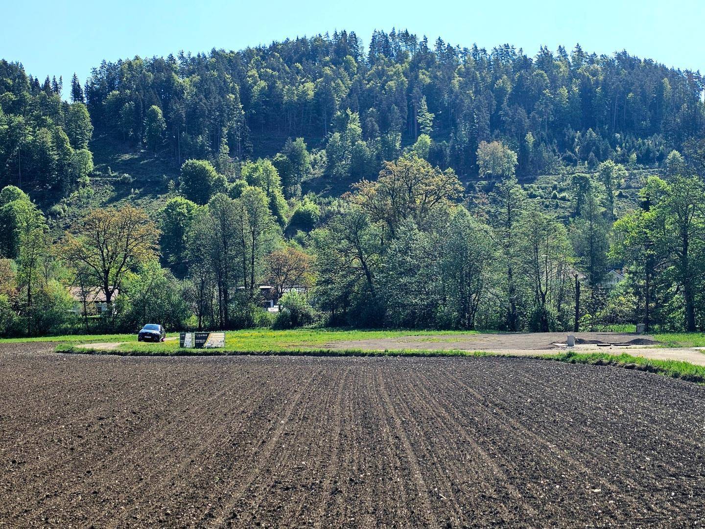 Weitläufiges gepflügtes Feld mit einem Auto in der Ferne und bewaldeten Hügeln im Hintergrund.