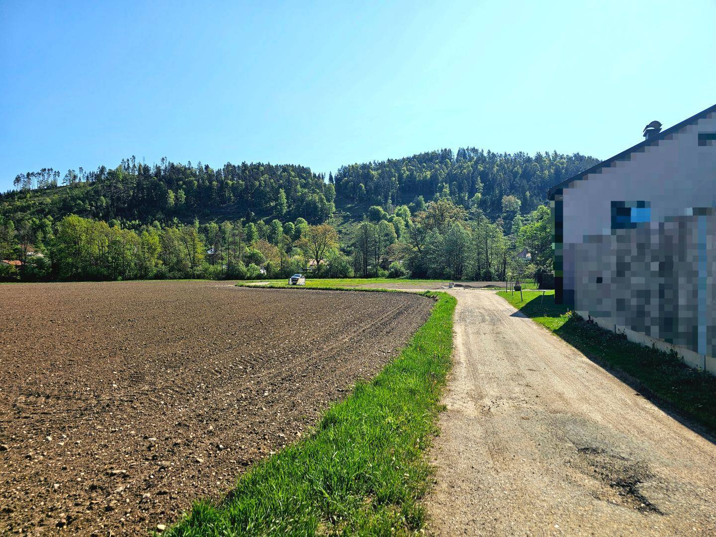 Gepflügtes Feld neben einem Feldweg mit üppigen bewaldeten Hügeln unter blauem Himmel.