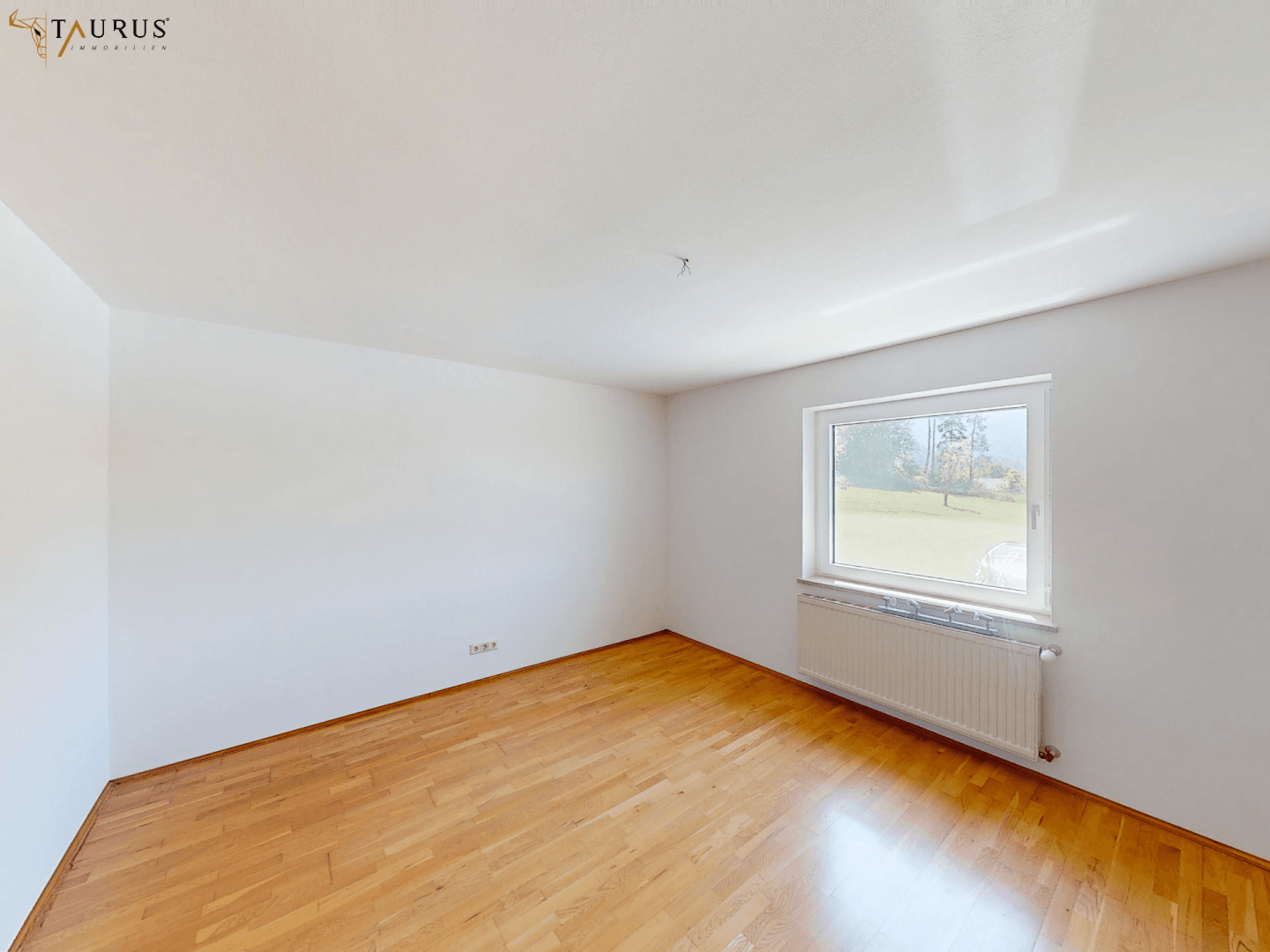 Helles Zimmer mit Parkettboden und einem Fenster, das den Blick ins Grüne freigibt.
