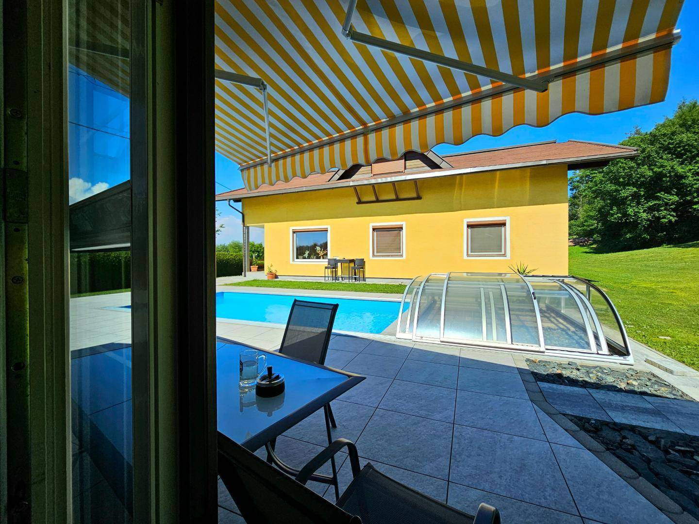 Blick von innen auf die sonnige Terrasse mit Markise und den erfrischenden Poolbereich.