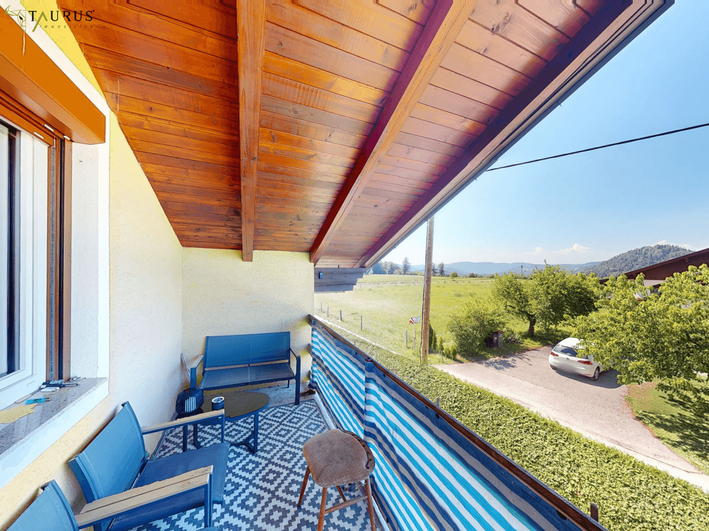 Sonniger Balkon mit Holzdach, Sitzgelegenheiten und weitem Blick über Felder und Berge.