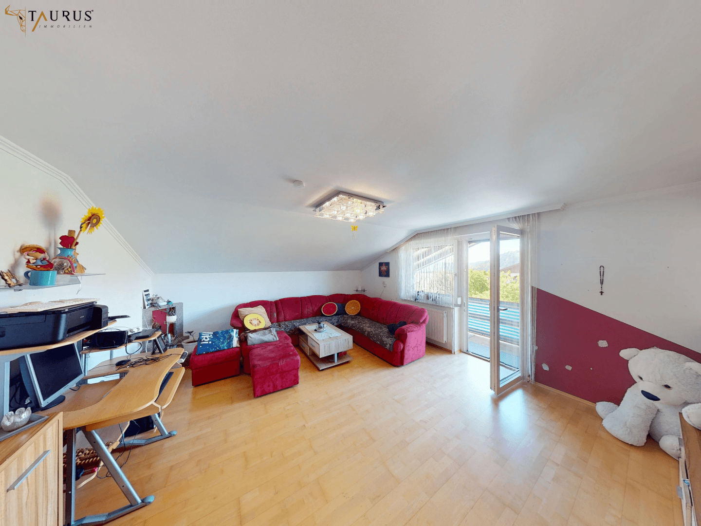 Helles Wohnzimmer mit rotem Ecksofa, Arbeitsbereich und Zugang zum Balkon.