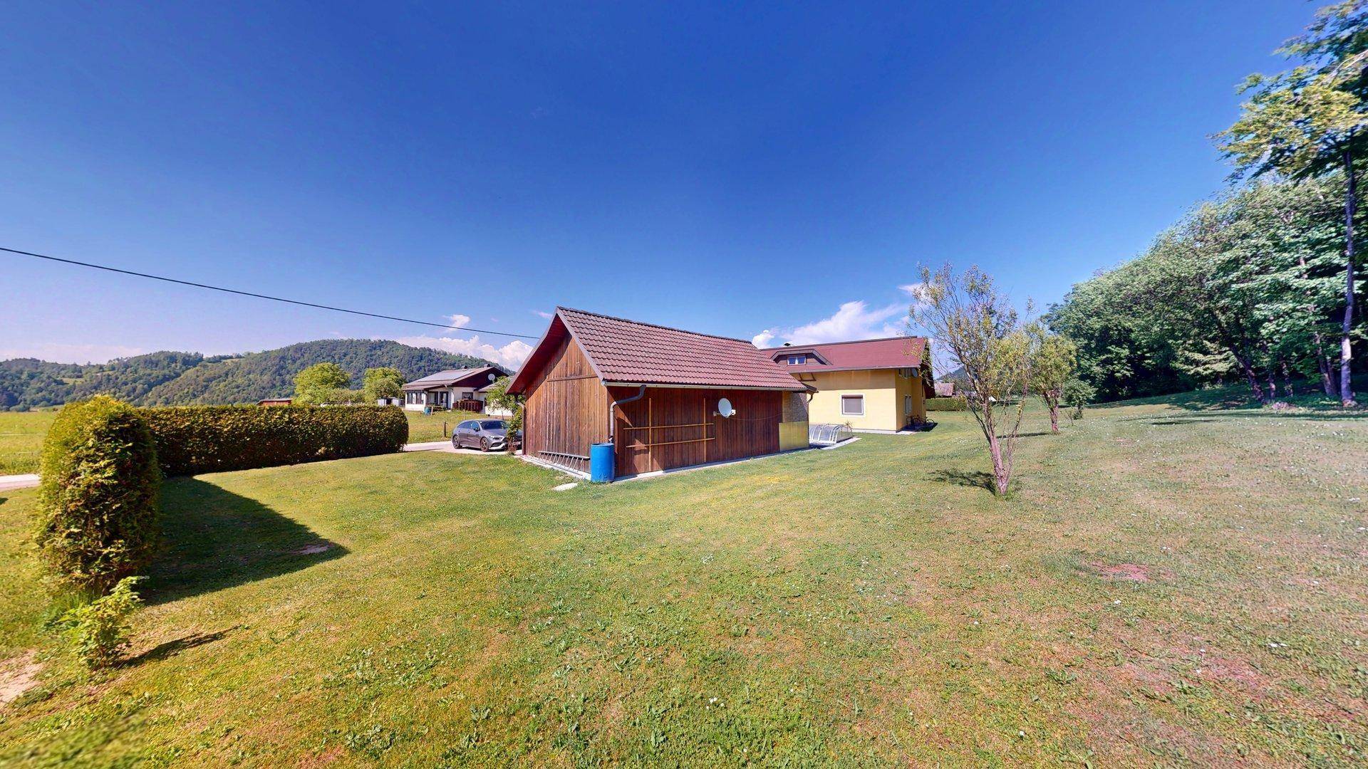 Das Holz-Nebengebäude mit Carport und das Haupthaus im Hintergrund, umgeben von Grünflächen.