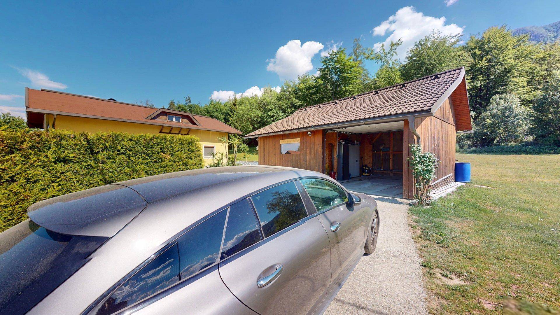 Ein Auto parkt vor dem Holz-Nebengebäude mit Carport, umgeben von Bäumen und Grünflächen.