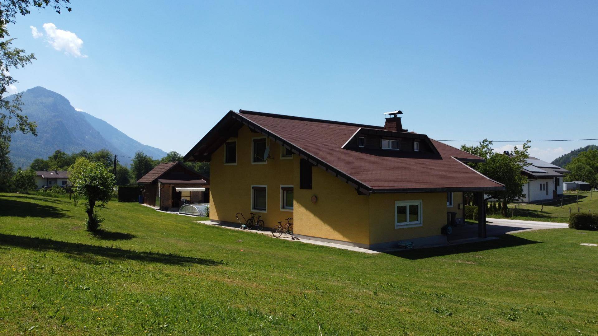 Seitenansicht des Hauses mit gelber Fassade, umgeben von viel Grün und Bergblick.