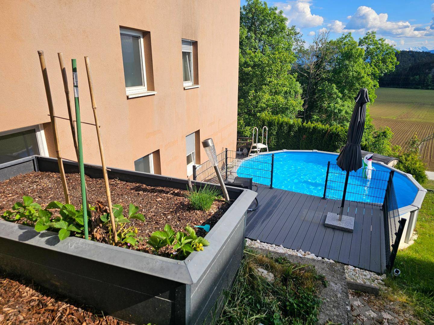 Seitenansicht des Hauses mit Hochbeeten und dem Poolbereich inklusive Sonnenschirm.