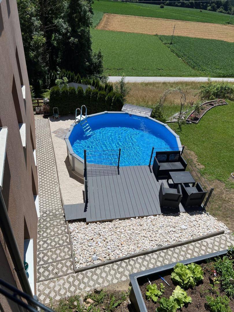 Gartenbereich mit Aufstellpool, Holzdeck und Sitzgelegenheiten, umgeben von grünen Feldern.