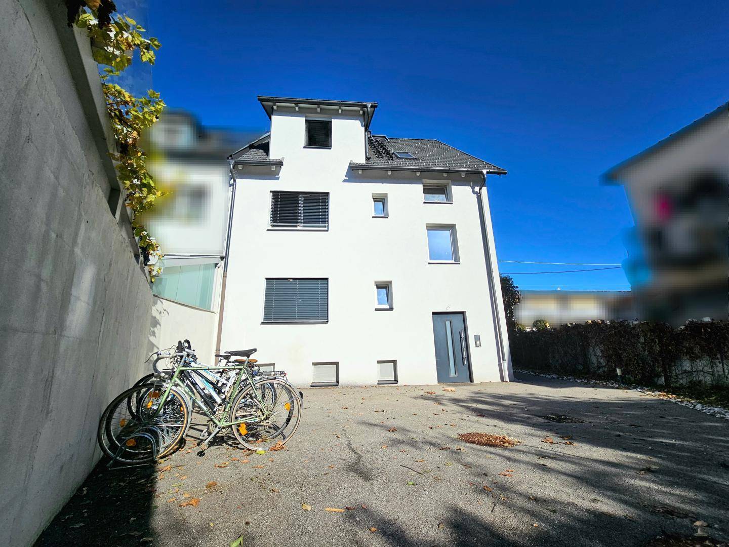 Weißes Wohnhaus mit Parkplatz und Fahrrädern an einer Mauer, unter blauem Himmel.