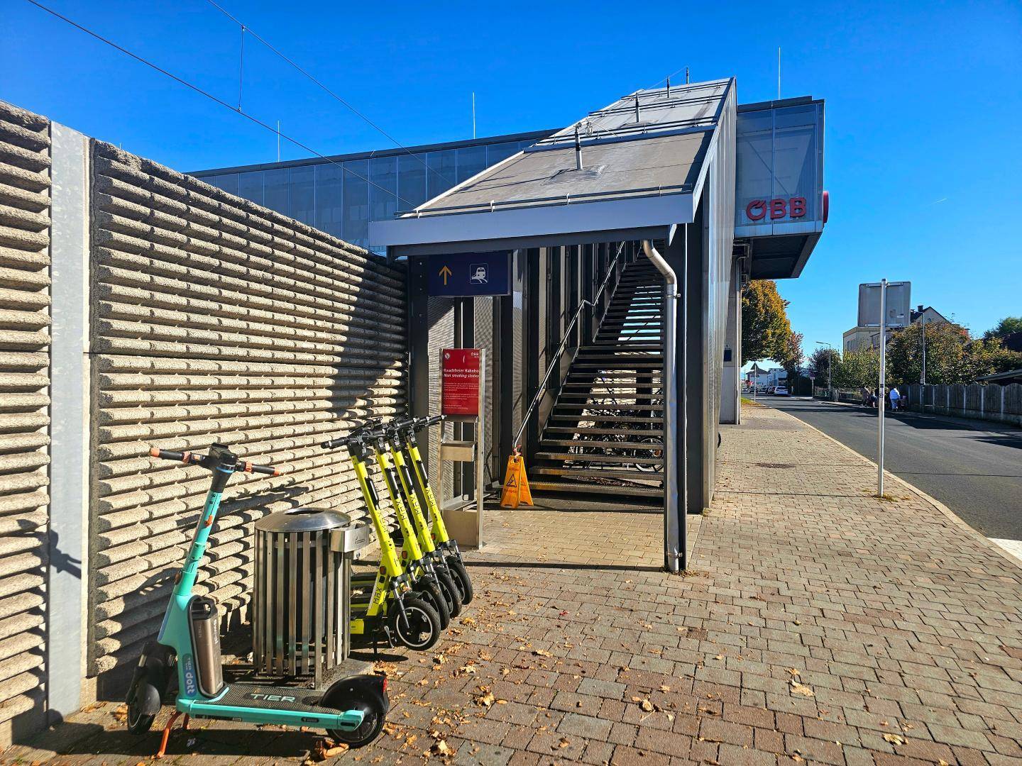 Außenbereich einer ÖBB-Haltestelle mit Treppenaufgang, Betonwand und abgestellten E-Scootern.