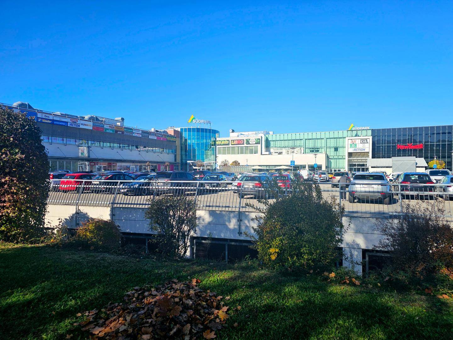 Großes Einkaufszentrum mit weitläufigem Parkplatz und umliegender Grünfläche unter blauem Himmel.