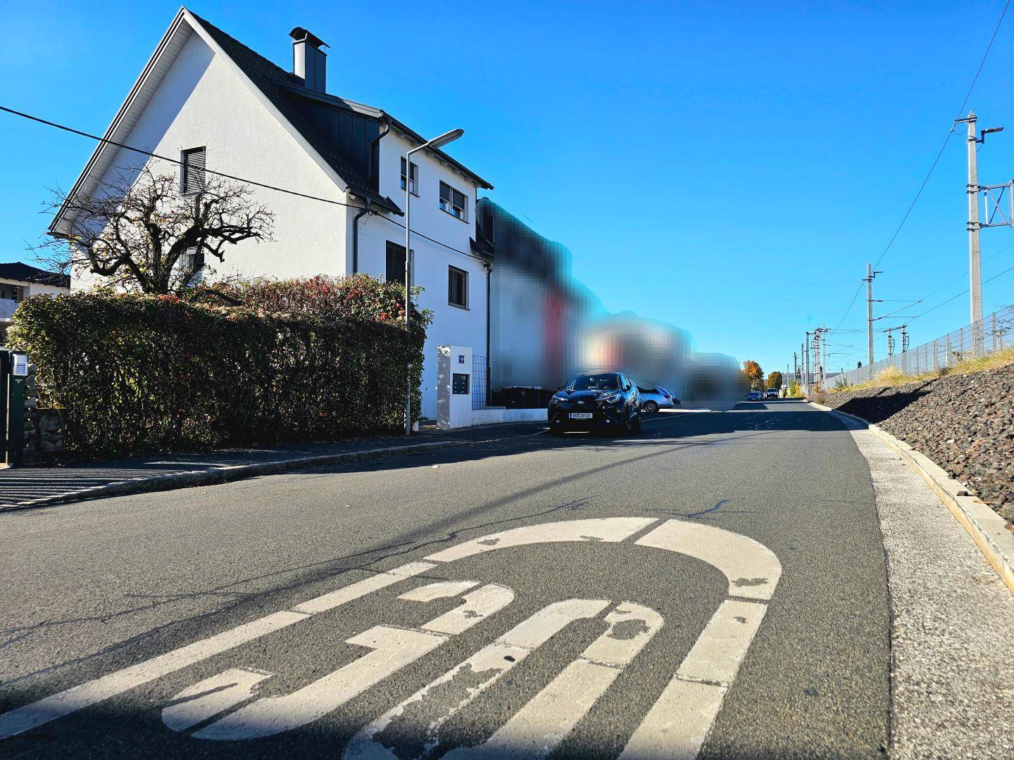 Weißes Wohnhaus an einer Straße mit Geschwindigkeitsbegrenzung und Hecke im Vordergrund.