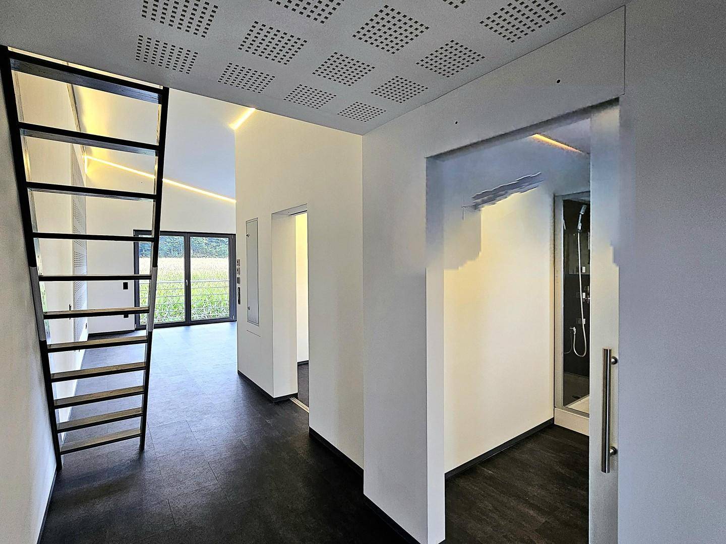 Heller Flur mit dunklem Boden, moderner Treppe und Blick auf große Fenster mit Grünblick.