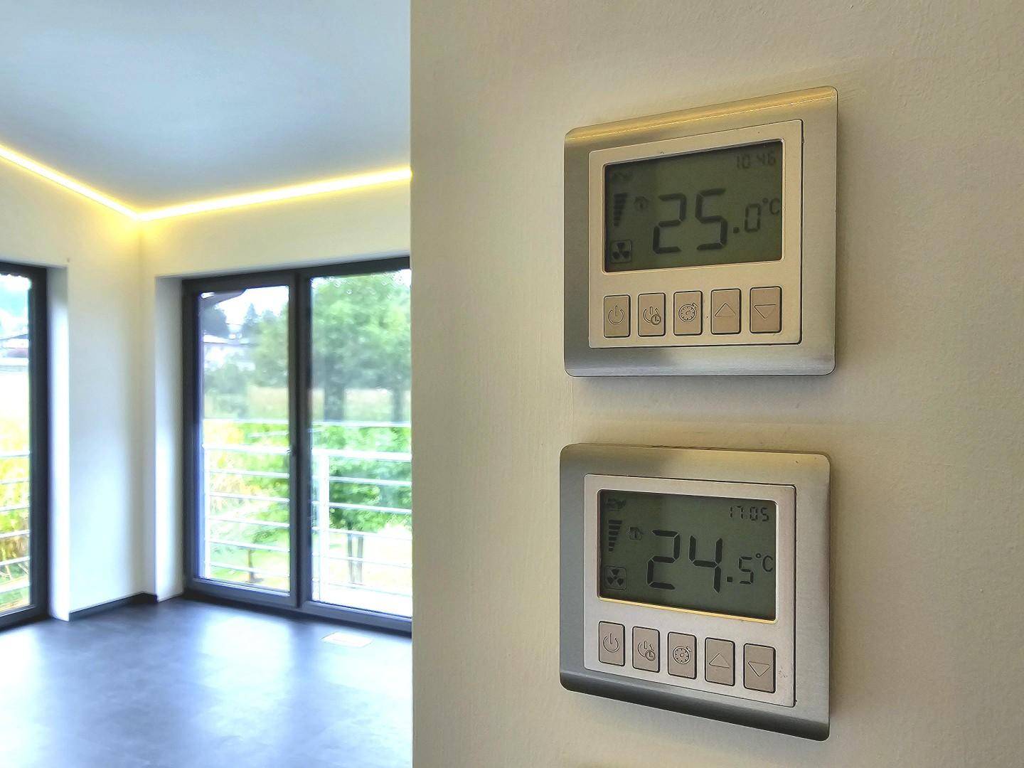 Zwei digitale Thermostate an einer weißen Wand mit Blick auf große Fenster.
