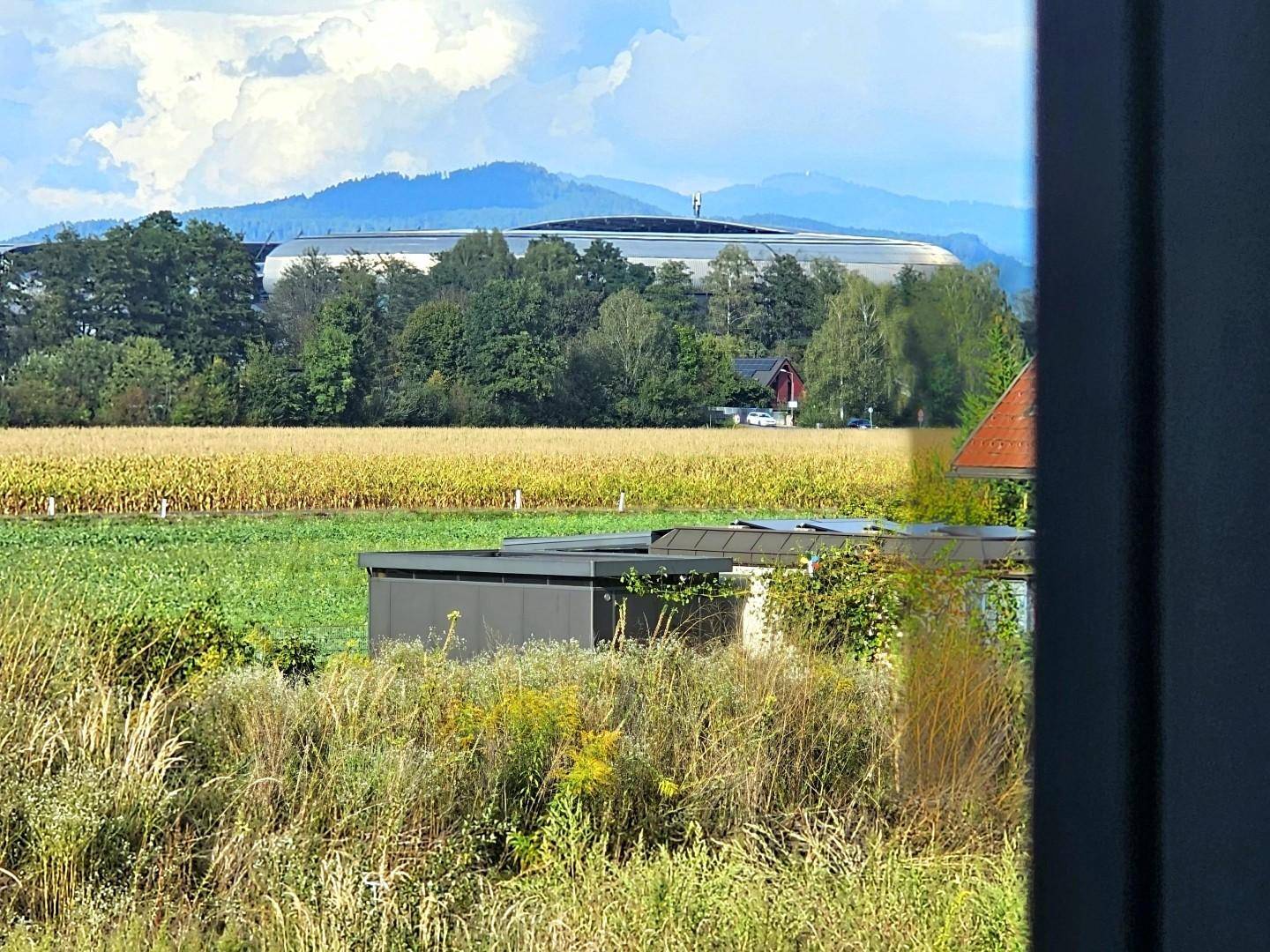 Weitläufiger Blick über Felder und Wiesen mit Bergen im Hintergrund und einem Stadion.