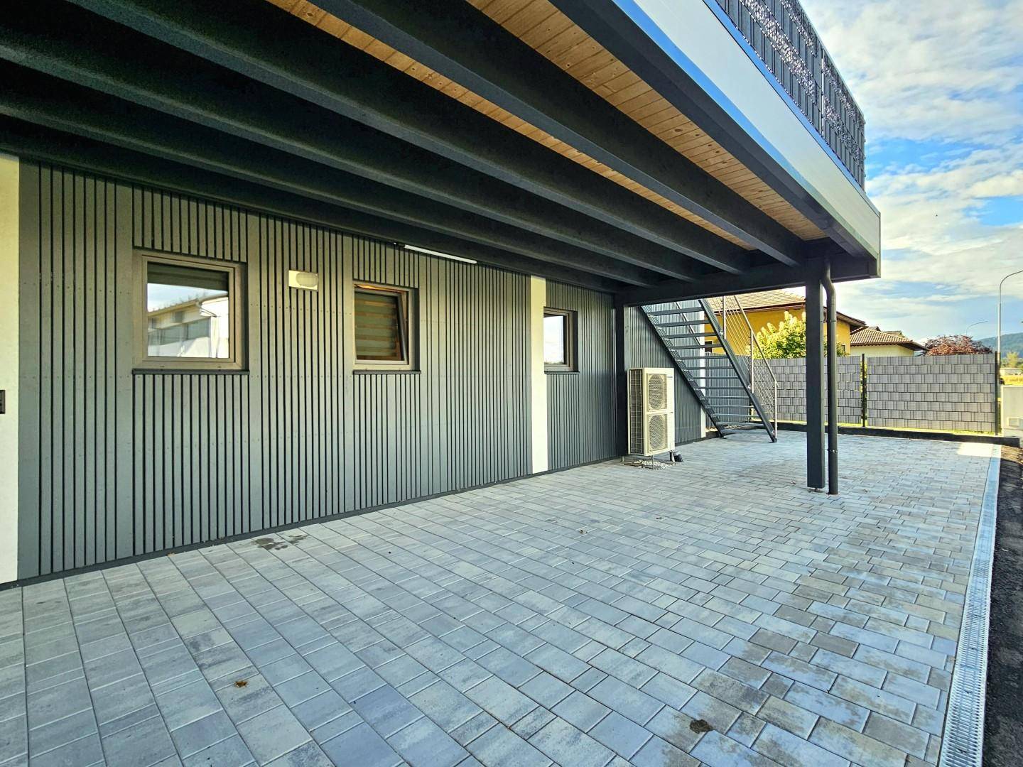 Überdachter Außenbereich mit Pflastersteinen, Außentreppe und Klimaanlage am Neubau.
