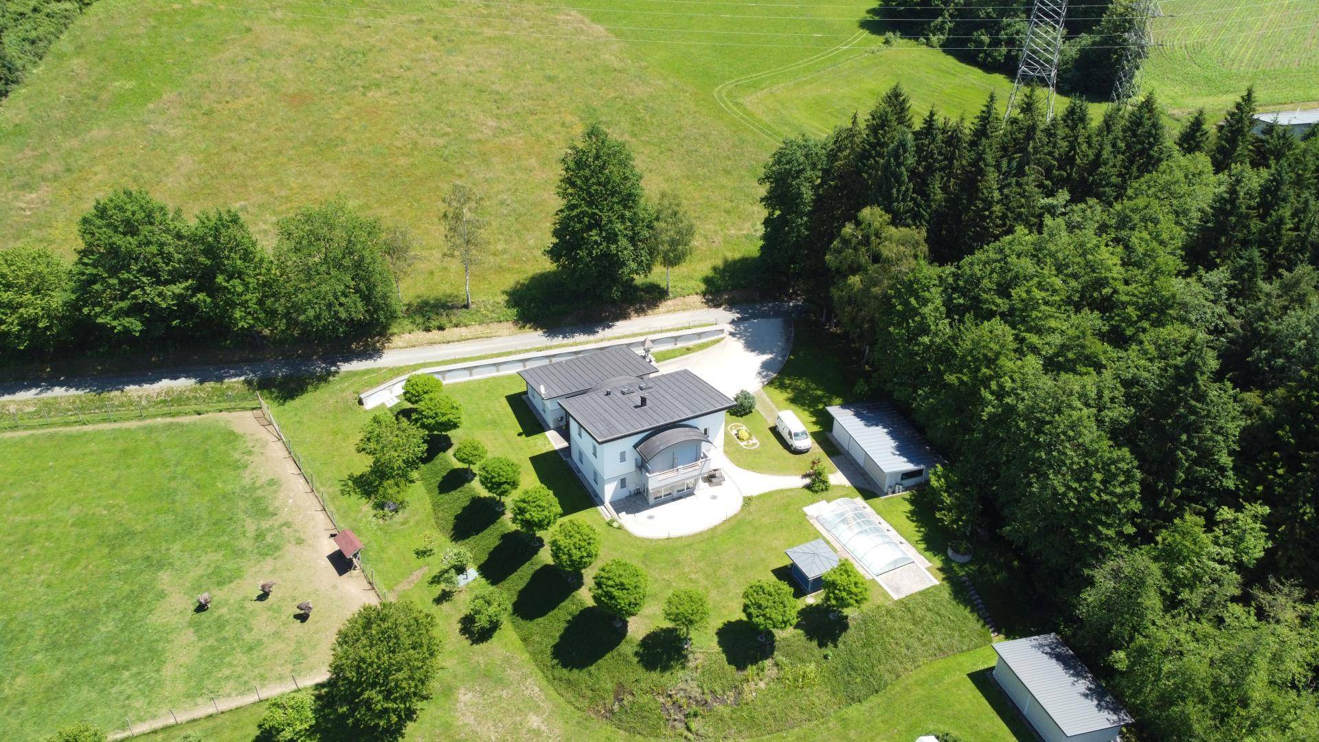 Luftaufnahme eines modernen Hauses umgeben von grünen Feldern und Wäldern unter blauem Himmel.
