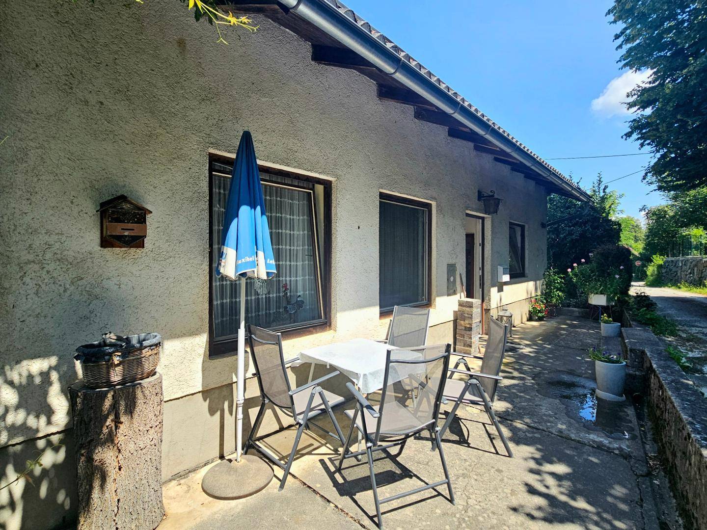 Sonniger Terrassenbereich mit Tisch, Stühlen und Sonnenschirm, umgeben von viel Grün.