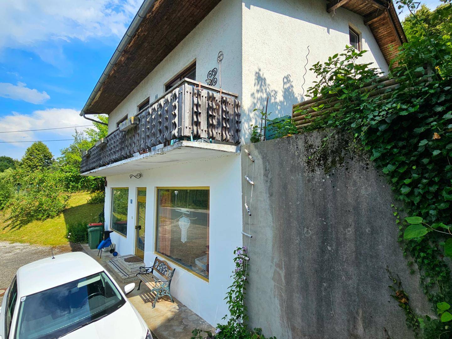 Seitenansicht des Hauses mit Balkon, Parkplatz und einer Betonmauer im Vordergrund.