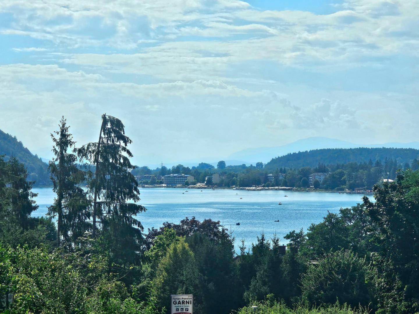 Panoramablick auf den blauen See, umgeben von grünen Hügeln und Wäldern unter bewölktem Himmel.