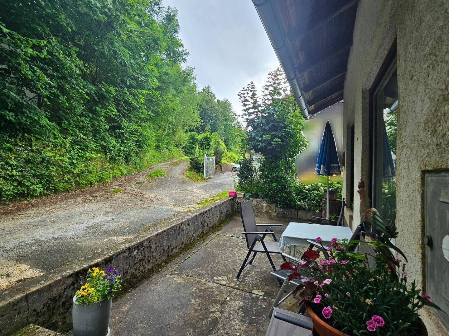 Terrasse mit Blumen und Gartenmöbeln, angrenzend an einen grünen Hang und einen Weg.