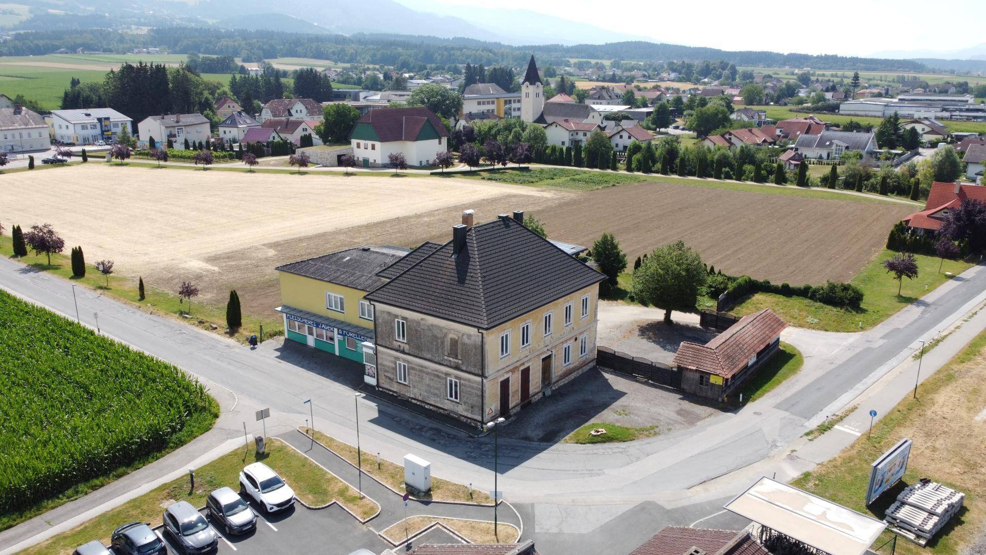 Drohnenansicht des Gebäudes mit angrenzenden Feldern und einem Dorf im Hintergrund unter blauem Himmel.