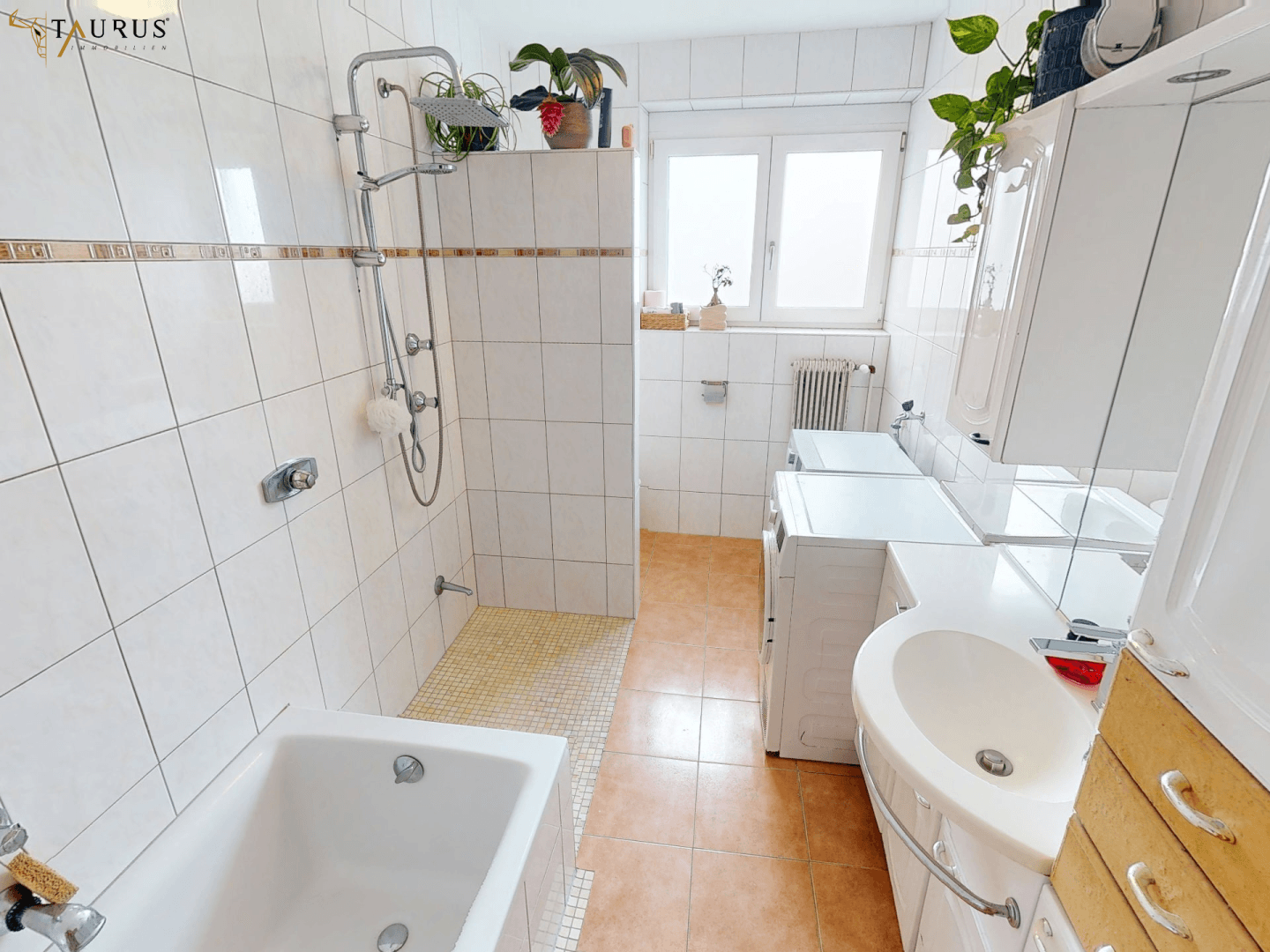 Gefliestes Badezimmer mit Badewanne, Dusche und Waschbecken, inklusive Waschmaschine und Fenster.